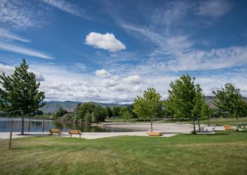 A wide shot of a brilliant blue sky with fluffy white clouds over a lawn, dotted with trees edging up against a sandy river shoreline. Low hills rise in the distance.