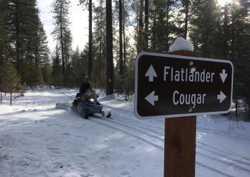 A person riding a snowmobile down a snow-covered path. A sign for Flatlander and Cougar trails is in the foreground.