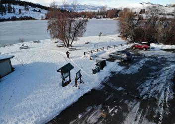 Plowed parking lot next to a snow-covered picnic area, overlooking a frozen lake with snow-covered mountains in the background.