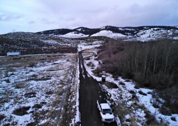 A plowed highway runs between a forest and a meadow with patchy snow with snowy hills in the background.