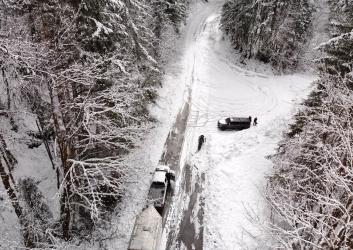Thick snow covers a pull-off parking area along a forest road surrounded by a snowy forest. A truck with a trailer sits on the forest road and another truck sits on the pull-off.