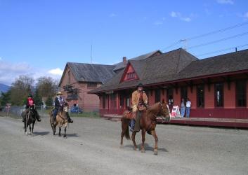 Three horse riders on brown, tan and black horses walk on a dirt road in front of an old trail stations painted orange with red trim. Two people wearing jeans and tshirts lean against the building watching the horse riders go by. 