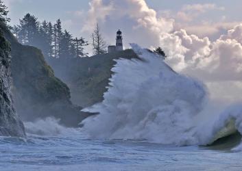 High king tide wave crashes against rocky cliffside with a lighthouse