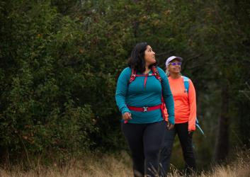 Two women, one in blue and one in orange, hike in the forest wearing backpacks.