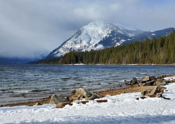 snow covered mountains in background above lake with snowy beach