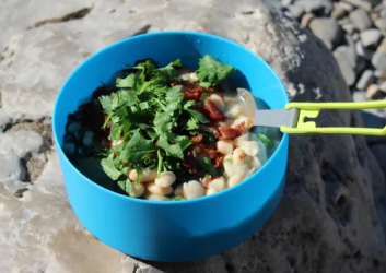 Blue bowl on a rock filled with beans, cilantro and bacon. A foldable spork is pressed into the mixture