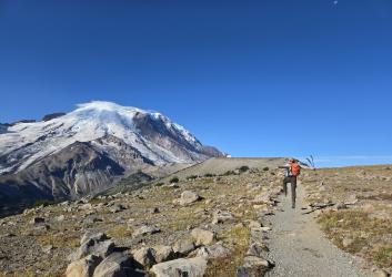 Man with hiking poles skips down a trail on a sunny day at Mt. Rainier National Park. There is a large mountain in front of him.