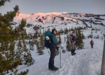 Two women snowshoe at sunrise below a snowy mountain.