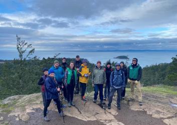 Group of hikers standing on a summit with evergreen trees and water in background