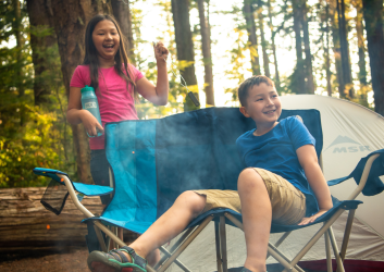 Boy sitting in a campchair with a girl behind him holding a roasting stick with a tent in the background
