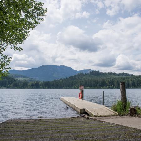 Water coming to a sloped boat launch next to dock with green evergreen pines across the lake and white clouds against blue sky.