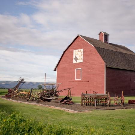 Red hey barn surrounded by early 1900 farm equipment. Green hills in the distance and light grey clouds against blue sky. 