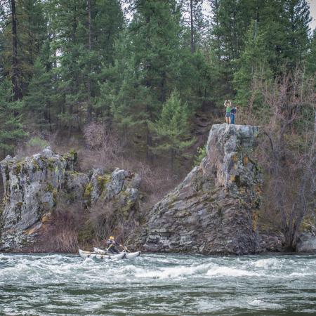 A boat rows through a river rapid while two visitors look on atop a cliff on the far side of the river.