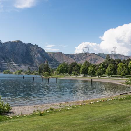 The grassy lawn gently slopes down to a narrow, curved beach with the calm water of the Columbia River and tall mountains in the background