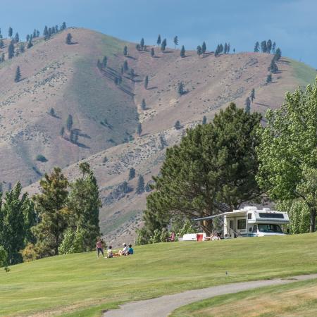A small RV sits atop a grassy hillside and four people picnic on the lawn. Tall trees and a large mountain are in the distance.