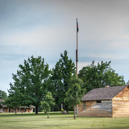 120 foot flag pole in the middle of the parade ground with large green trees and log cabins close by.