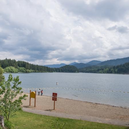 Sandy swim beach area with no lifeguard sign. Bench facing the water play area, Evergreen pinetrees surrounds the lake.