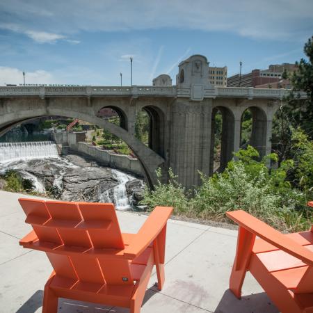 Two chairs sit along the edge of the Centennial Trail. A waterfall is visible underneath a large concrete bridge.