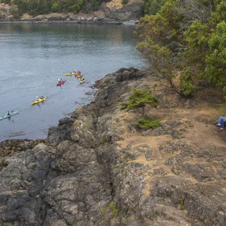 A group of six kayaks paddle along the shoreline of the rocky cliff as two hikers take a rest at a picnic table to watch the kayakers pass by. There are green trees and shrubs  around the onlookers.