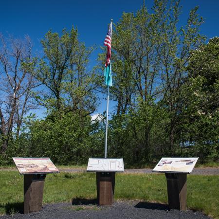 Three interpretive panels sit in front of a flag pole with the American and Washington State flags in front of a line of trees and a blue sky