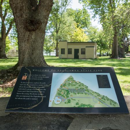 A welcome plaque with the park map sits on a rock foundation in front of a historic tan building. The plaque and building are shaded by the trees and surrounded by grass.