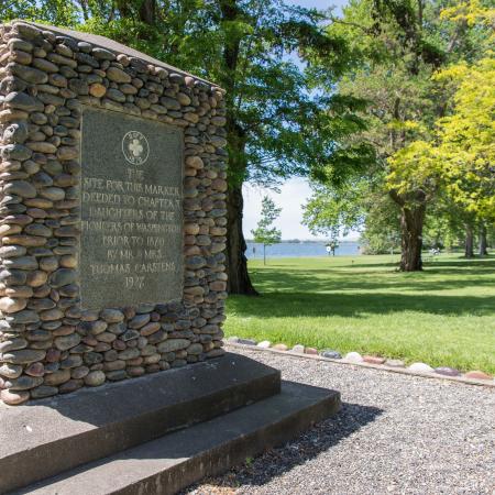 A rock monument from the Daughters of the Pioneers of Washington is set in a gravel pad with green grass and shade trees around the pad.  The river can be seen in the distance.