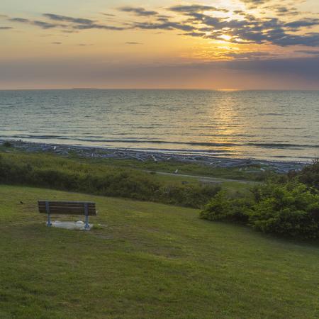 A lone bench sits in a grassy lawn looking over the water as the sun sets, creating a blue, orange and pink sky with some clouds covering the sun. 