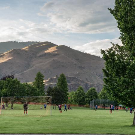 Brown hills with few trees are in the background with the cloudy blue sky. In the foreground, soccer is being played on green grass with trees around the soccer field edges. 