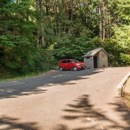 A small red car is parked in an asphalt paved parking lot, lined with cement bumper blocks. A brown restroom sits behind the red car. A sign on the left side of the parking area reads, "No overnight parking or camping" with a Discover Pass required sign underneath. Tall evergreen trees surround the entire area. 