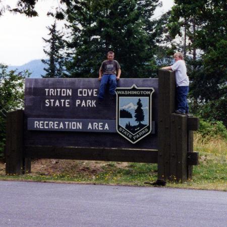 Two children sit atop a wooden sign reading Triton Cove State Park Recreation Area and bearing the Washington State Parks Logo shield. Pine trees rise in the background.