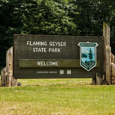 A wooden sign reading Flaming Geyser State Park Welcome and bearing the Washington State Parks Shield