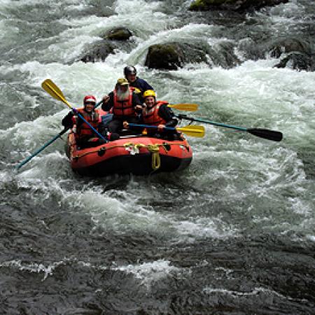 Four people in water safety gear and helmets aboard an orange and black river raft and holding yellow paddles float past rocks on river whitewater.