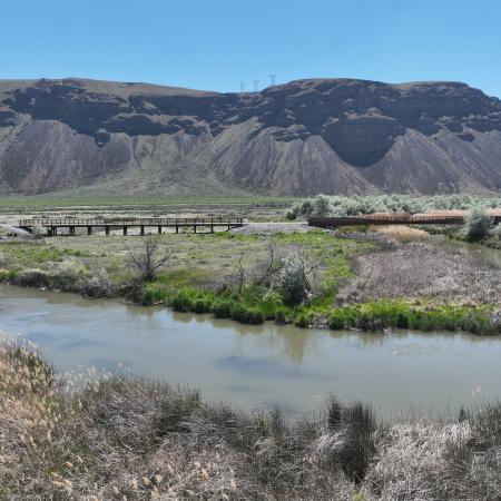 Large basalt hill and blue sky overlooking two trestles, one over water and one over land connecting a straight trail. 