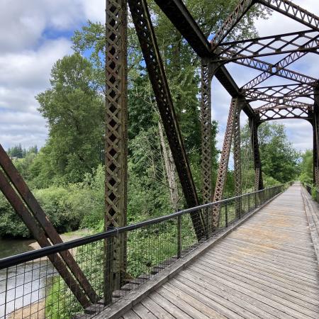 Looking down an old iron train trestle bridge over a river that has been converted to a bike path. It has a wooden deck and is surrounded by green trees and is under a blue sky.