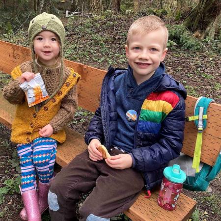 Young girl and boy bundled in warm jackets with First Day Hike buttons sitting on park bench eating snacks