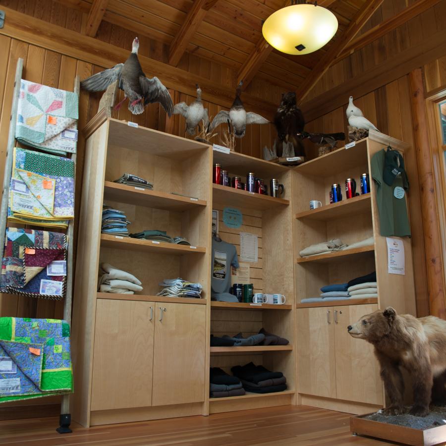 Inside the contact station looking at the items available for purchase. Items include quilts, shirts, mugs, water bottles, and coffee thermoses. There are 6 taxidermy birds visible as well as a taxidermy bear cub.
