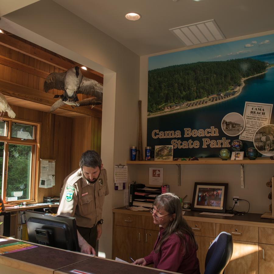 Two employees working at the contact station where Discover passes can be purchased and you can check-into cabins. The building is rustic with lots of exposed wood. There are some taxidermy birds handing from the ceiling. Three are visible from this angle. 