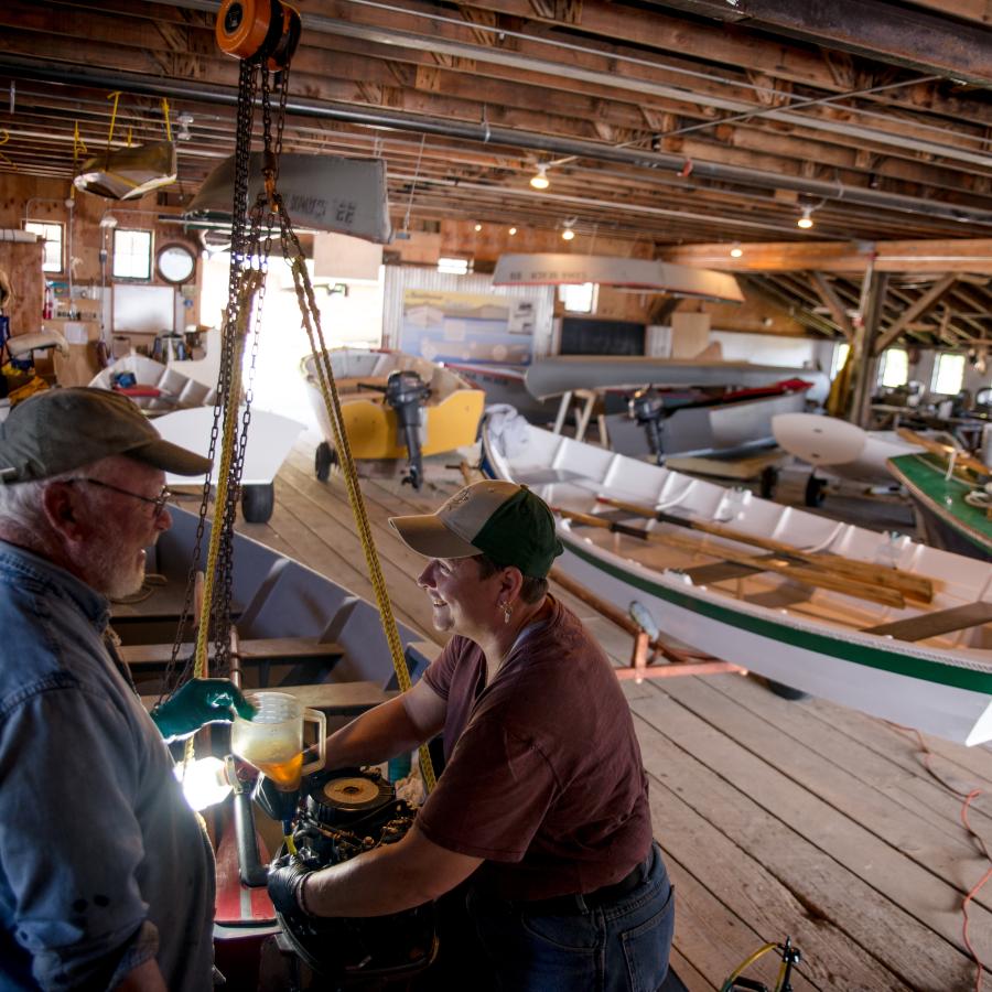 Inside the boat center is two people in ball caps who appear to be working on one of the wooden boats. There are at least 10 boats visible in the photo. Many are white, but there is a visible yellow and green boat.  The inside of the building has exposed wooden and metal rafters and multiple visible lights. 