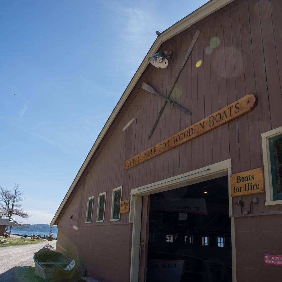 The boat center at Cama Beach is a large barn-like building with dark brown wooden siding. A wooden sign says "the center for wooden boats." Another says "boats for hire." There are some other signs, but they are difficult to make out. There are two decorative oars on the building. 