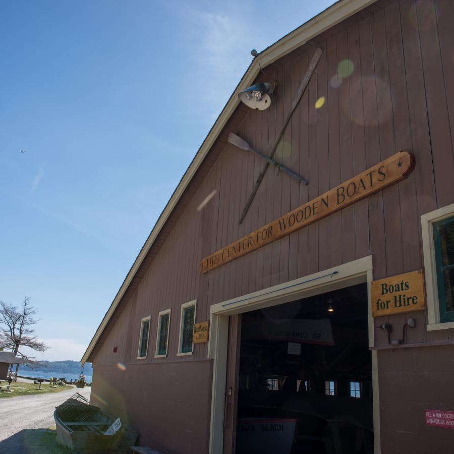 The boat center at Cama Beach is a large barn-like building with dark brown wooden siding. A wooden sign says "the center for wooden boats." Another says "boats for hire." There are some other signs, but they are difficult to make out. There are two decorative oars on the building.