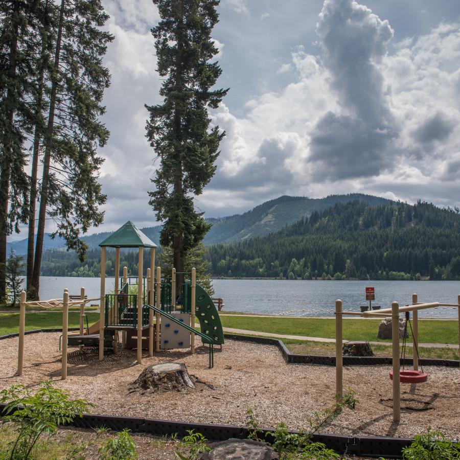 An outside child play structure with adjacent tire swing sitting on play bark. Located off to the side of the green grass behind the swimming area. Dark green evergreens across the lake and white clouds against a blue sky.