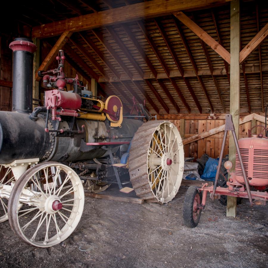 A coal powered tractor and an early model gas powered tractor on display underneath a covered wood shed.