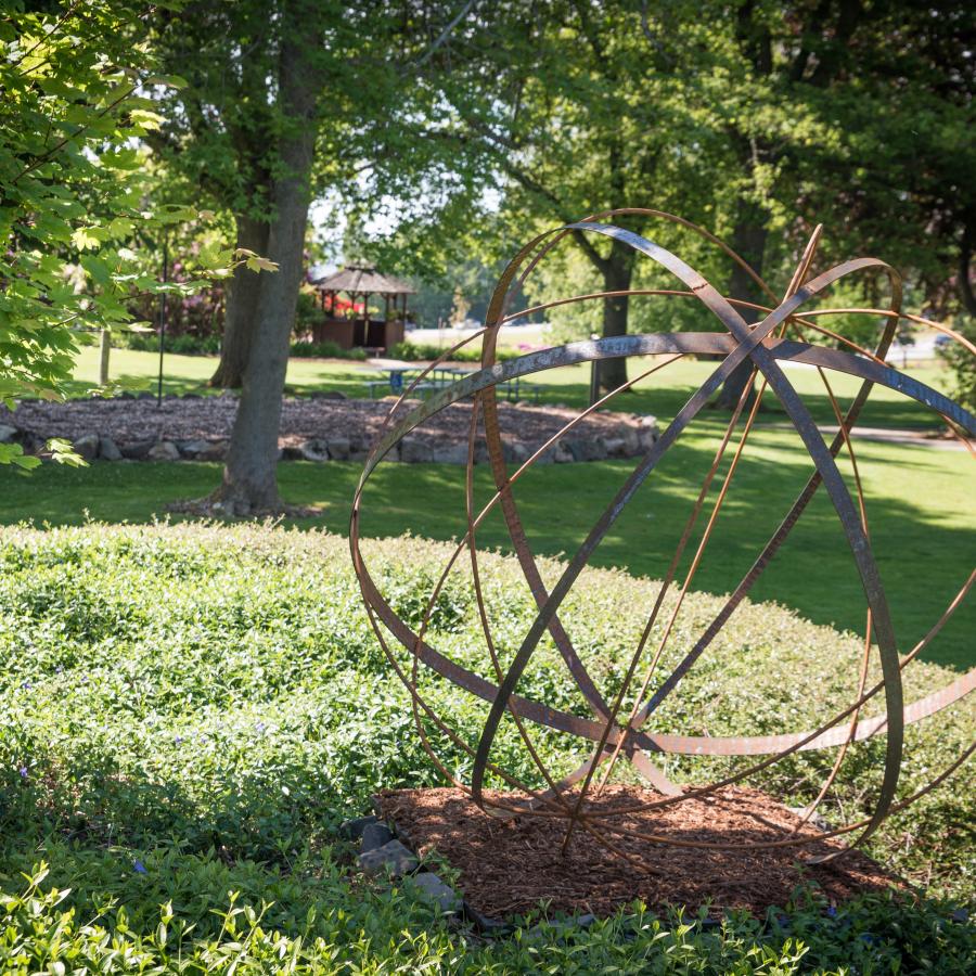 Circular metal art piece in the park on a sunny day surrounded by green grass and bushes
