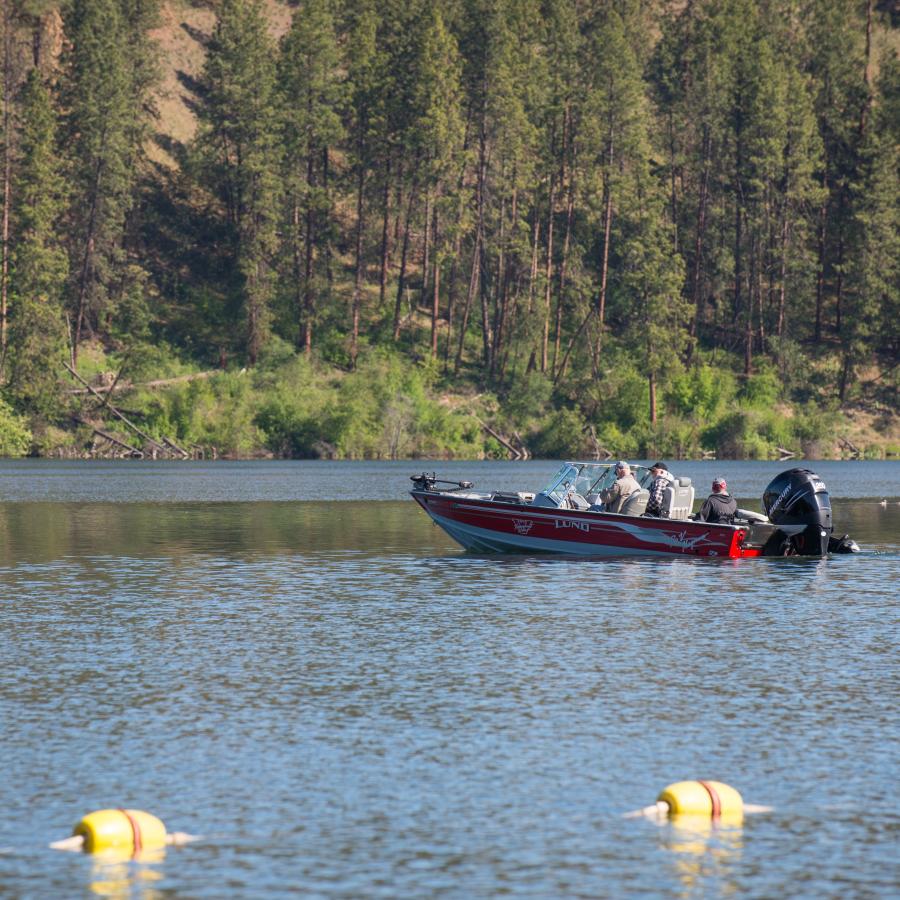 Three visitors in a red motorboat on a lake with the edge of a swimming area in the foreground and trees and bushes on the far side of the lake.