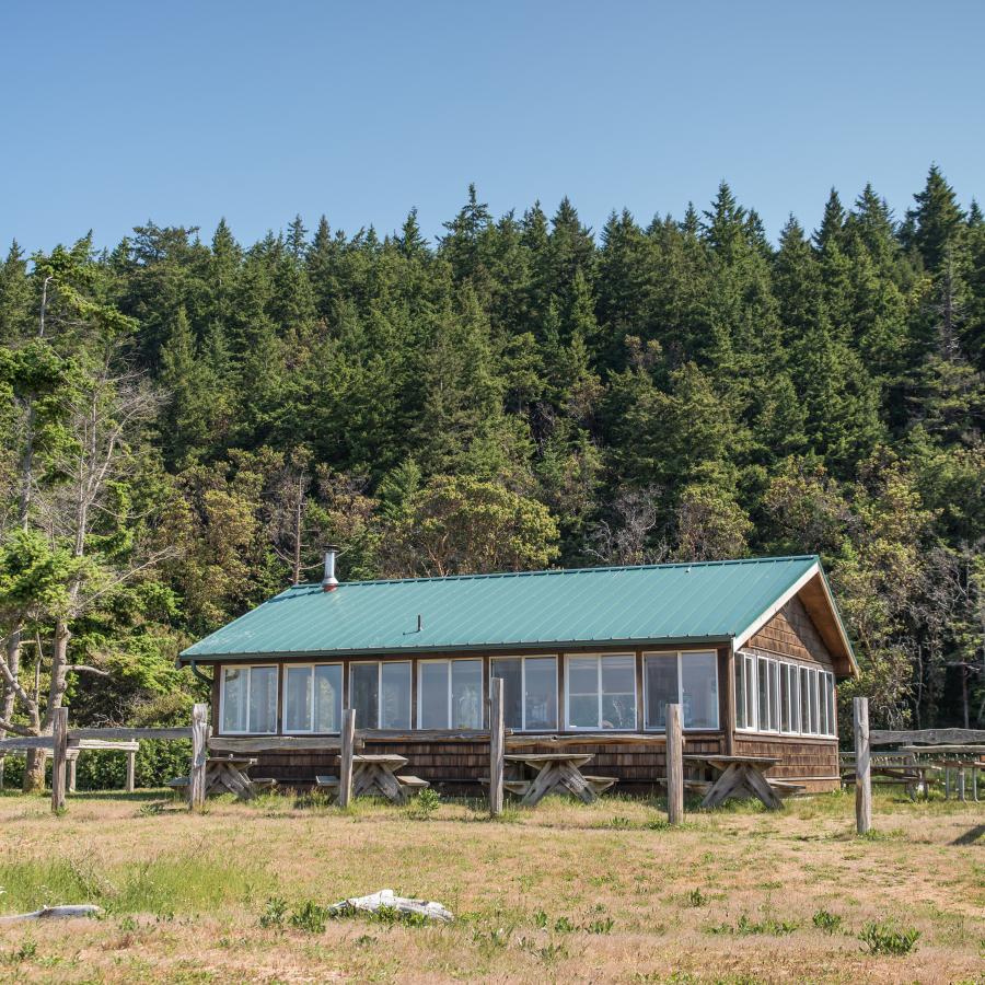 Back and left side of the picnic shelter exterior. The back of the picnic shelter has 7, large, white windows that fill the entire back side. Along the left side of the building there is 5 white-framed windows which span the entire left side of the building.  The roof is metal-looking and green and the siding is a dark brown stained wood. There is a wood post-fence visible with some of the fencing materials missing so that all you see is posts. There are picnic tables visible along both sides. 