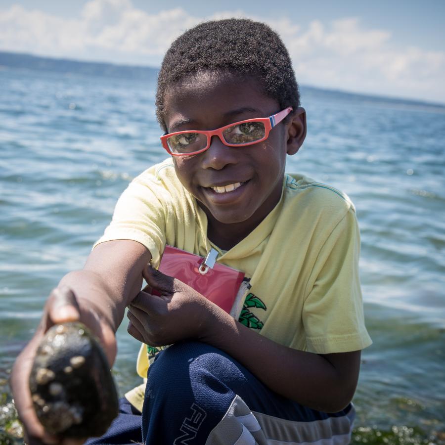 Smiling, black youth with a green shirt, blue pants, and red glasses displaying a rock they found on the beach.  The rocky beach and Saratoga Pass are visible in the photo but are blurred.