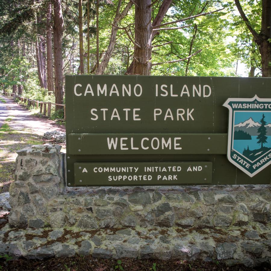 Entrance sign at Camano Island State Park. Trees are visible in the background as well as a road on the left hand side. 