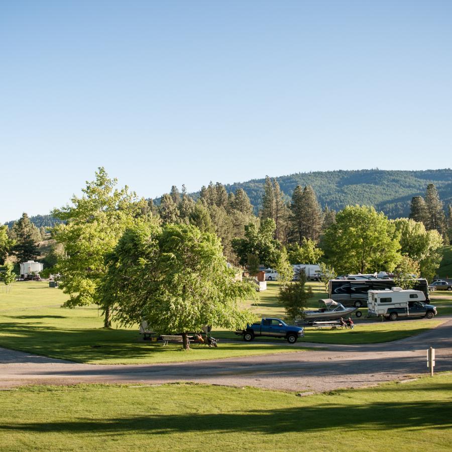 A road runs through a flat, grassy campground with intermittent trees, RVs, and hills in the distance.