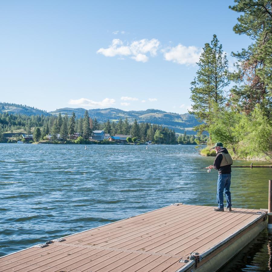 A gentleman stands at the end of a dock fishing into the lake.