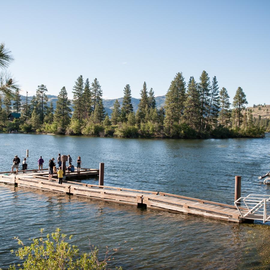 A group of people stand at the end of a dock fishing into the lake with tall trees and bushes along the far shore.